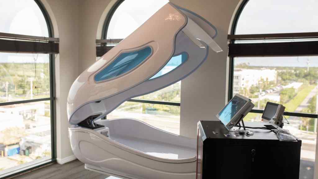 A white, futuristic medical or wellness pod with an open lid is positioned in a room with large arched windows, overlooking a cityscape. The setting feels modern and serene.