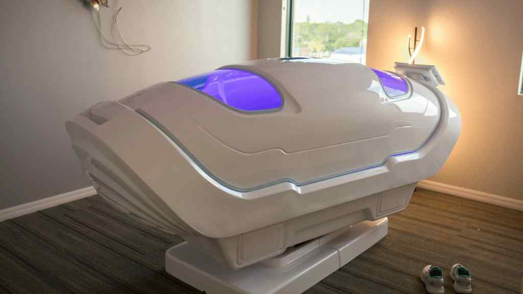 A sleek, futuristic white pod with blue-tinted windows sits in a softly lit room. Wires are attached to the wall, and a pair of shoes are placed nearby, suggesting possible human interaction or rest.